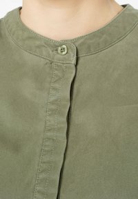 Olivengrühes Hemd mit rundem Kragen, Knopfverschluss und strukturiertem Stoff. Verfügt über kontrastierende Nähte entlang der Knopfleiste.