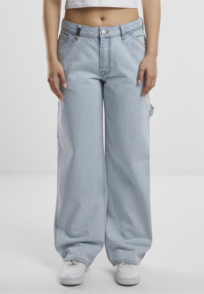 Osoba u svijetloplavim širokim trapericama i bijelim tenisicama stoji pred jednostavnom svijetlom pozadinom.