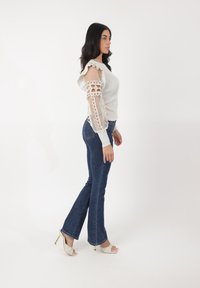 Maglione grigio chiaro a coste con scollatura a volant e maniche lunghe trasparenti e ricamate. Jeans a zampa blu scuro con vita media e sandali con tacco beige.