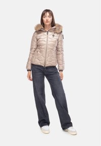 Giacca imbottita beige con cappuccio e bordo in pelliccia sintetica, motivo di cuciture diagonali, chiusura con zip e tasche laterali, abbinata a jeans scuri.
