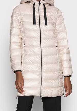 Lange, hellbeige gesteppte Steppjacke mit schwarzem Reißverschluss, schwarzen Kordeln an der Kapuze und seitlichen Reißverschlusstaschen, getragen über einer schwarzen Hose.