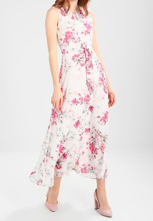 Femme portant une robe maxi blanche sans manches avec un imprimé floral rose et des chaussures à talons hauts roses, debout devant un fond uni.