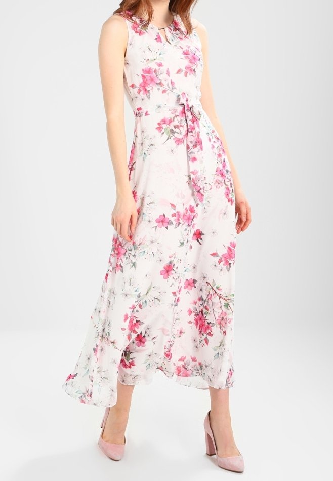 Femme portant une robe maxi blanche sans manches avec un imprimé floral rose et des chaussures à talons hauts roses, debout devant un fond uni.