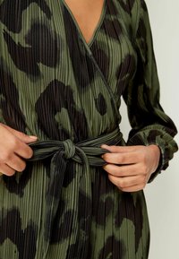 Vestido envolvente plisado en verde y negro con escote en v, mangas largas y cintura ajustada con lazo, que presenta un sutil patrón de estampado animal.