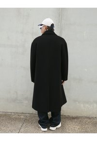 Manteau long noir avec grands revers et silhouette droite, associé à un pantalon en denim bleu et des baskets blanches, vu de l'arrière.
