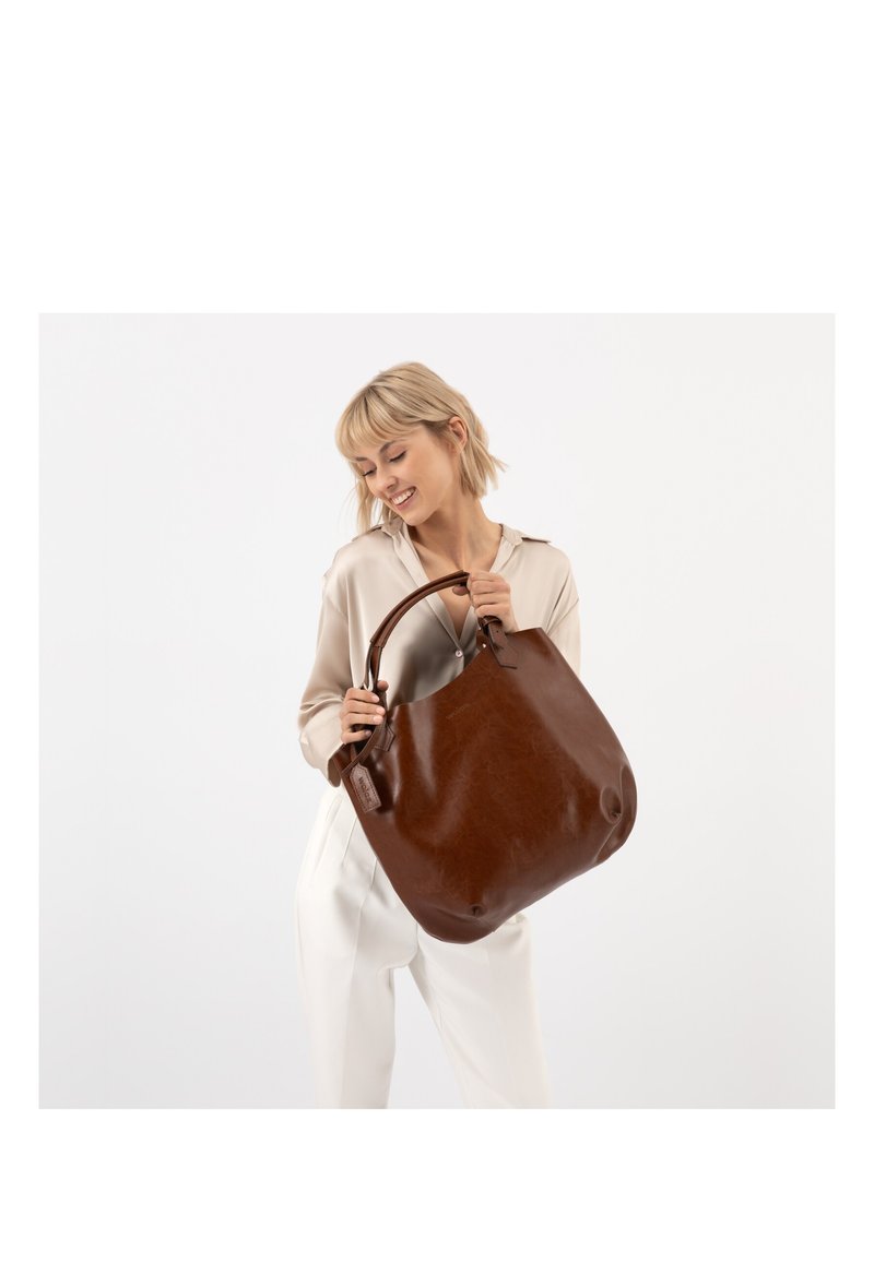 Femme souriante aux cheveux blonds courts tenant un grand sac à main en cuir marron, portant un chemisier beige et un pantalon blanc sur fond blanc.
