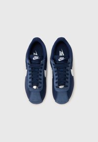 Sapatilhas azul marinho com uma mistura de materiais em camurça e rede, apresentando o símbolo da Nike em branco, atacadores e detalhes em metal. Design com bico arredondado.