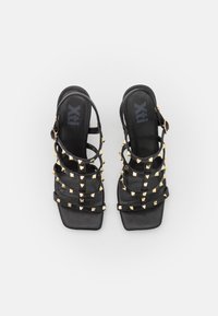 Sandalias de tacón alto negras con tiras, que presentan acentos de tachuelas doradas, punta rectangular y una delgada correa de tobillo asegurada con una hebilla de tono dorado.