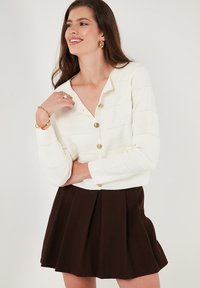 Weißer, verkürzter Cardigan mit strukturierten Streifen und goldenen Knöpfen, kombiniert mit einem braunen, plissierten Rock. Glattes Material und taillierte Passform.