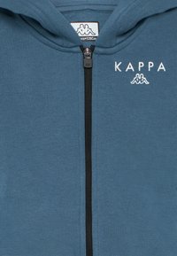 Zils kapuci svārsts ar priekšējo rāvējslēdzēju, ar izšūtu "KAPPA" logo un auduma tekstūru, ideāli piemērots ikdienas valkāšanai.