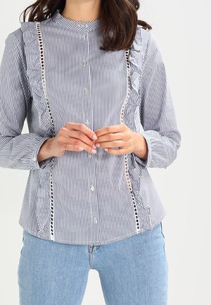 Kobieta w niebiesko-białej, pionowo paskowanej koszuli zapinanej na guziki z falbankami i koronkowymi detalami, w zestawie z jasnoniebieskimi dżinsami.