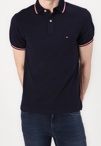 Polo-shirt bleu marine avec col et poignets rayés rouge et blanc, arborant un petit logo sur la poitrine. Tissu en coton, design ajusté.