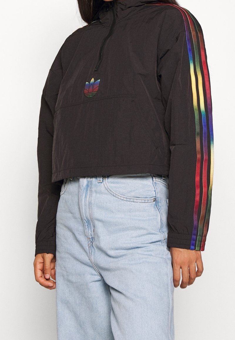 Giacca cropped nera con cappuccio, caratterizzata da maniche a righe arcobaleno e un logo colorato. Realizzata in materiale leggero, texture liscia.