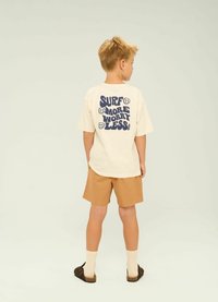 Camiseta beige con texto gráfico en azul marino "SURF MORE Worry LESS" y caras sonrientes. Combinada con pantalones cortos beige y zapatos oscuros tipo slip-on. Fondo claro.