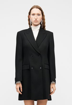 Kvinde med langt bølget hår iført en sort dobbeltradet blazer-kjole over en hvid rullekrave og lange dråbeformede krystaløreringe.