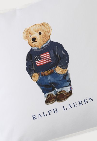 Λούτρινο αρκουδάκι που φοράει μπλε πουλόβερ με σημαία των Ηνωμένων Πολιτειών, μπλε τζιν, καφέ παπούτσια και ζώνη, με το κείμενο "RALPH LAUREN" από κάτω.