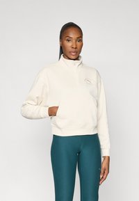 Pull-over zippé à quart en polaire crème avec une poche avant et un logo brodé. Associé à des leggings teal. Texture lisse, coupe décontractée.