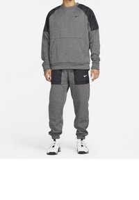 Homme portant un survêtement Nike gris et noir avec des baskets blanches et noires, debout sur un fond blanc.