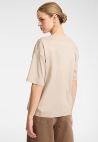 T-shirt in cotone beige con spalle scese, maniche corte e vestibilità rilassata. Il tessuto ha una texture liscia e un design semplice.