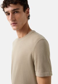 Pull court en tricot beige à manches courtes avec un col rond et des manches retroussées. Tissu texturé avec un motif horizontal subtil.