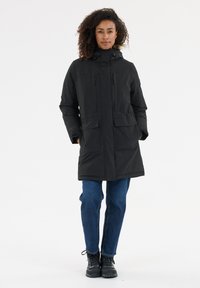 Zwarte pufferjack met capuchon, rits aan de voorkant, twee grote voorkezen en verstelbare trekkoorden. Gecombineerd met blauwe jeans en zwarte laarzen.