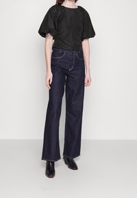 Top nero a maniche puff con scollatura rotonda, abbinato a jeans a gamba larga blu scuro. Le scarpe sono nere con dettagli rosa sui tacchi.