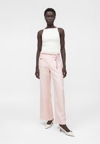 Femme debout portant un haut blanc sans manches, un pantalon large rose clair avec poches avant, et des escarpins blancs pointus avec des nœuds.