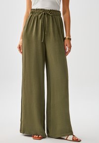 Pantaloni a gamba larga color verde oliva realizzati in tessuto strutturato con vita elastica regolabile con coulisse e spacchi laterali, abbinati a sandali color cuoio.