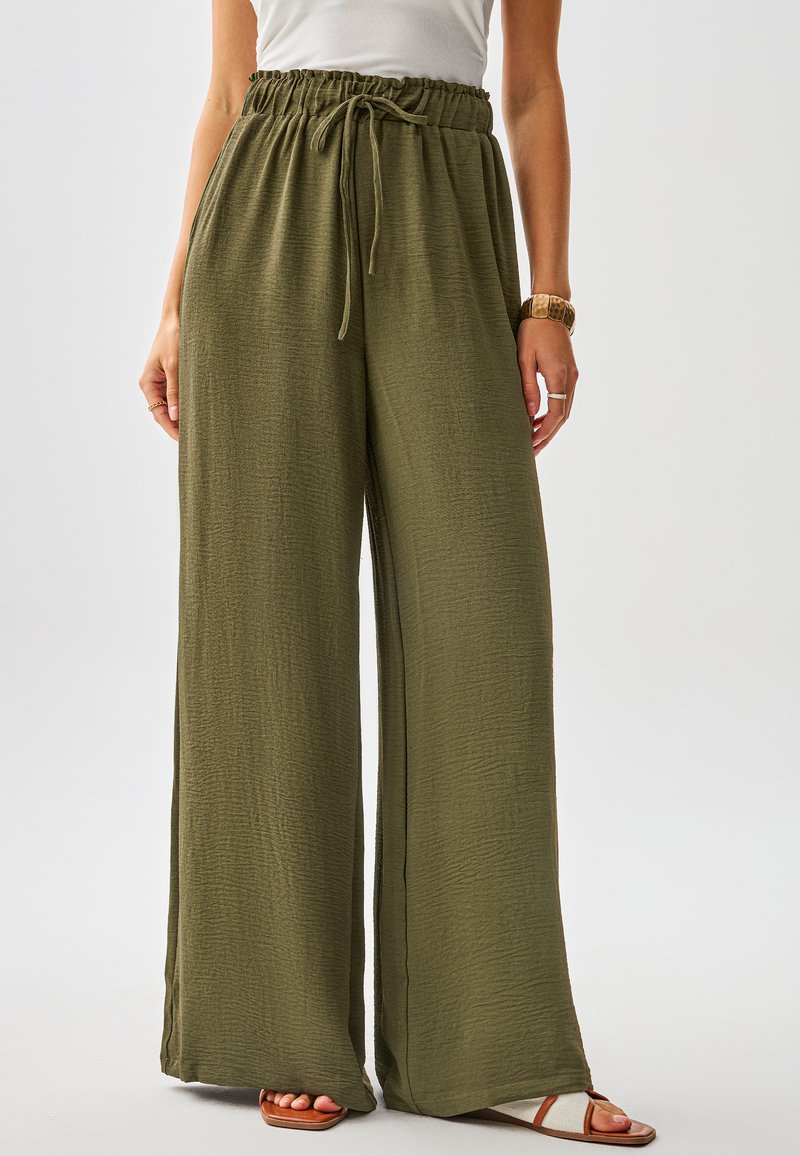 Pantaloni a gamba larga color verde oliva realizzati in tessuto strutturato con vita elastica regolabile con coulisse e spacchi laterali, abbinati a sandali color cuoio.