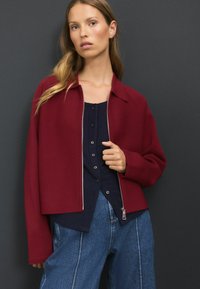 Veste bordeaux avec un col et une fermeture éclair sur le devant, associée à un haut bleu marine à boutons et un jean en denim, offrant une coupe décontractée et une texture lisse.