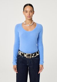 Vrouw in een lichtblauw fluffy langemouwen shirt, bredere donkere jeans, een riem met koeienprint en een chunky parelketting, recht naar voren kijkend.