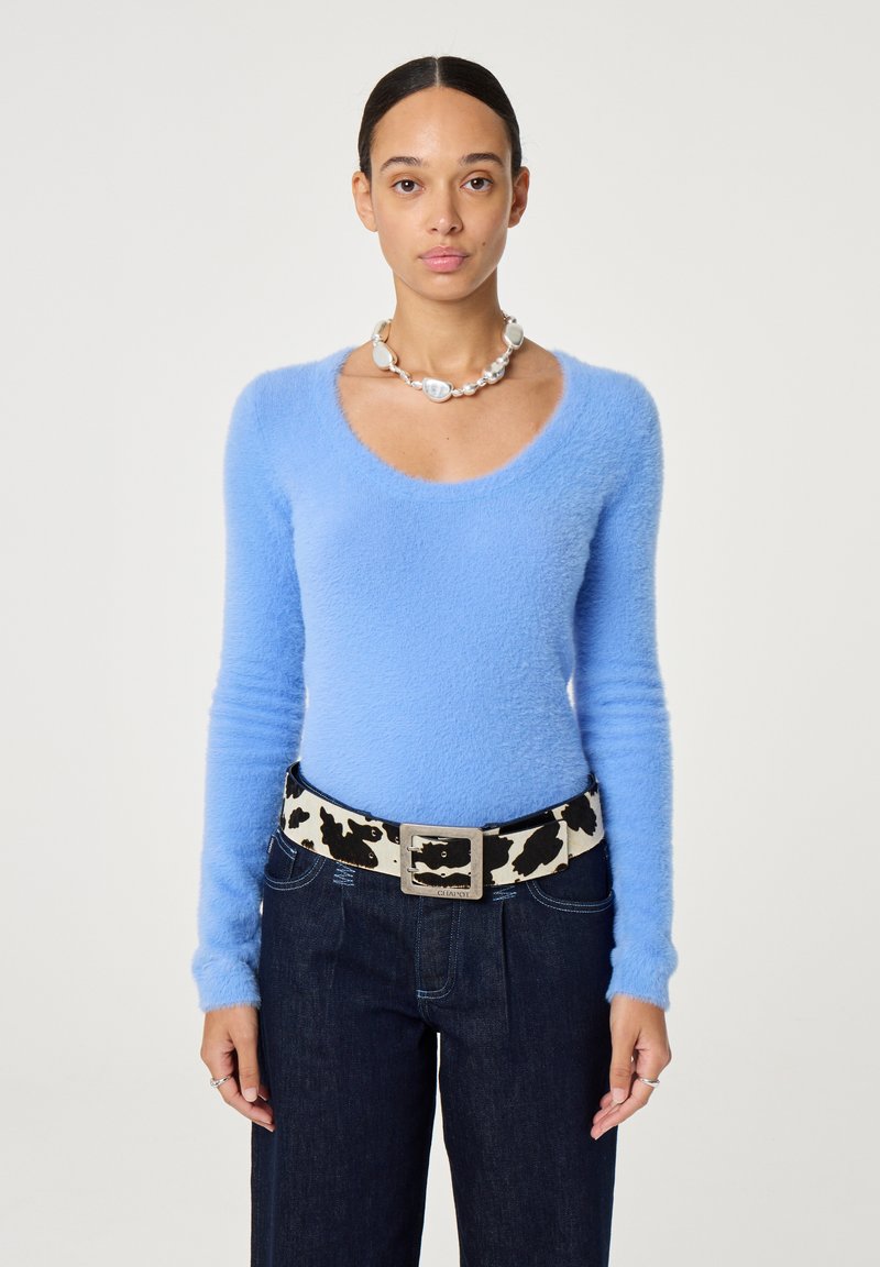 Vrouw in een lichtblauw fluffy langemouwen shirt, bredere donkere jeans, een riem met koeienprint en een chunky parelketting, recht naar voren kijkend.
