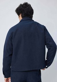 Veste bleu marine au design structuré, texture lisse et col légèrement rehaussé. Présente des poignets boutonnés et des coutures ouvertes.