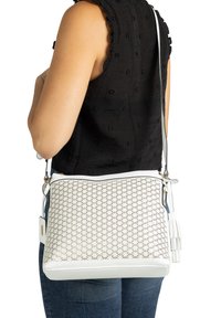 Frau trägt ein schwarzes Oberteil und Jeans, hält eine weiße Umhängetasche mit floralem Cutout-Design und Quastenakzent am Riemen.