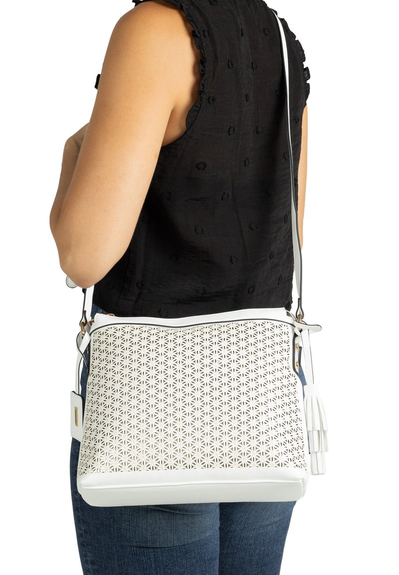Frau trägt ein schwarzes Oberteil und Jeans, hält eine weiße Umhängetasche mit floralem Cutout-Design und Quastenakzent am Riemen.