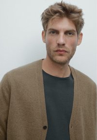 Cardigan di lana marrone con scollatura a V e chiusura con bottoni, indossato sopra una maglietta grigia scura. La texture è morbida e calda.