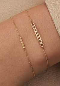Bracelet en ton doré présentant deux chaînes distinctes : l'une avec un charme en barre plate et l'autre avec des maillons entrelacés. Texture lisse, design minimaliste.