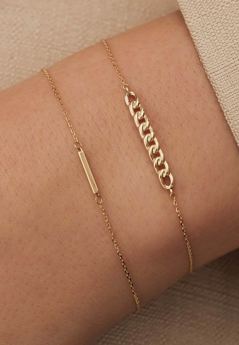 Bracelet en ton doré présentant deux chaînes distinctes : l'une avec un charme en barre plate et l'autre avec des maillons entrelacés. Texture lisse, design minimaliste.