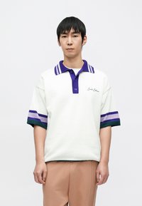 Biała koszulka polo o fakturowanym wyglądzie, z fioletowo-zielonymi paskami na rękawach i kontrastującym kołnierzykiem. Haft z napisem "Carlo C.".