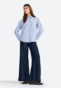 Camicia a righe blu con dettagli arricciati sul davanti, abbinata a pantaloni in denim scuri a gamba larga con orlo sfilacciato.