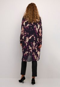 Abito lungo e trasparente in un motivo tie-dye viola e crema, abbinato a pantaloni neri aderenti e tacchi neri. Tessuto morbido e design fluido.