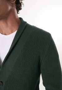 Tume roheline kudumismehhanismiga kardigan, millel on shawl krae, ribakujundus ja üks nööp sulgemiseks. Kantud valge T-särgi peal.