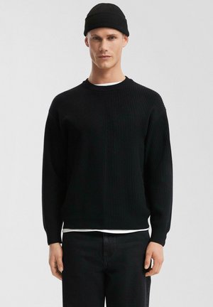 Männlicher trägt schwarzen Strickpullover, schwarze Mütze und schwarze Hose, steht vor einem schlichten hellgrauen Hintergrund.