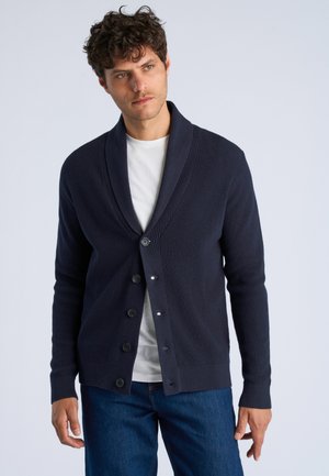 Man met krullend haar draagt een donkerblauwe sjaalkraagcardigan over een wit T-shirt en blauwe jeans tegen een effen achtergrond.