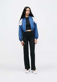 Blouson bomber court bleu et blanc porté sur un haut noir, associé à un pantalon noir taille haute et des chaussures blanches slip-on. Tissu lisse, coupe sportive.