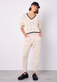 Beige gestrickter Pullover mit schwarzem Besatz, V-Ausschnitt und strukturiertem Muster; kombiniert mit hellbeigen schmal geschnittenen Hosen und schwarzen Plateauschuhen.