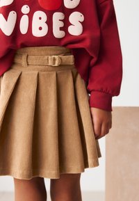 Jupe en velours côtelé beige avec plis et ceinture à boucle, associée à un sweat-shirt rouge avec un texte blanc audacieux. Texture lisse, design décontracté.