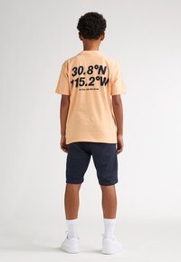 T-shirt pêche clair avec grand texte noir au dos, associé à un short bleu marine foncé et des baskets blanches. Manches courtes et coupe décontractée.