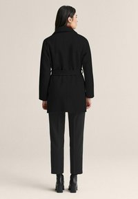 Musta vyöllä varustettu takki, jossa on räätälöity muotoilu, pitkät hihat ja lovikaulus. Päällä mustat housut ja korkokengät.