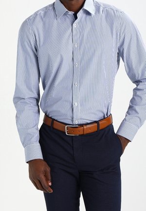 Mann trägt ein blau-weiß gestreiftes Hemd, in dunkelblaue Hose mit einem hellbraunen Ledergürtel gesteckt, eine Hand ruht in der Tasche.